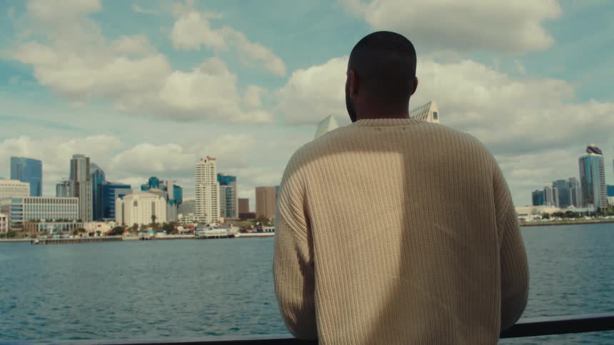 A young black man standing on a boat watching the city skyline in the distance during a sunny day boat ride
