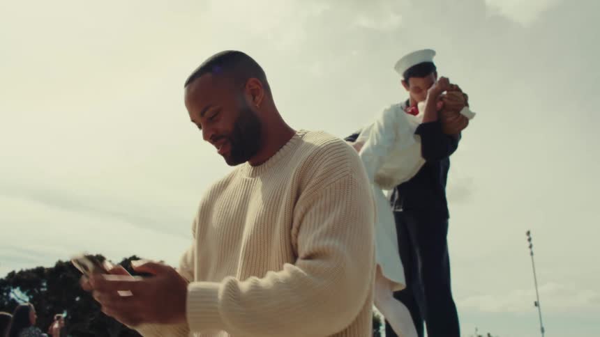 A man taking a selfie in front of a sailor statue kissing a woman at midday