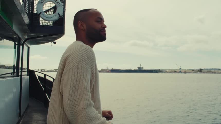 A medium shot shows a man wearing a cream sweater leaning against a railing on a boat as he looks out over the water toward a distant shoreline under a cloudy sky