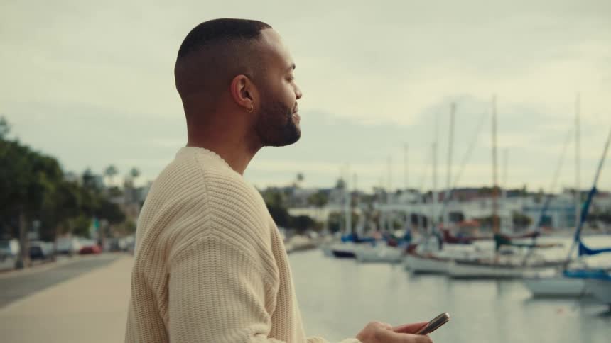 A medium side view shows a man in a cream sweater using a phone while standing on a walkway near a harbor filled with sailboats under a cloudy sky