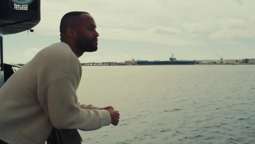 A man in a white sweater leans against a boat railing and looks at the ocean while a large naval ship and a distant shoreline sit under a cloudy sky