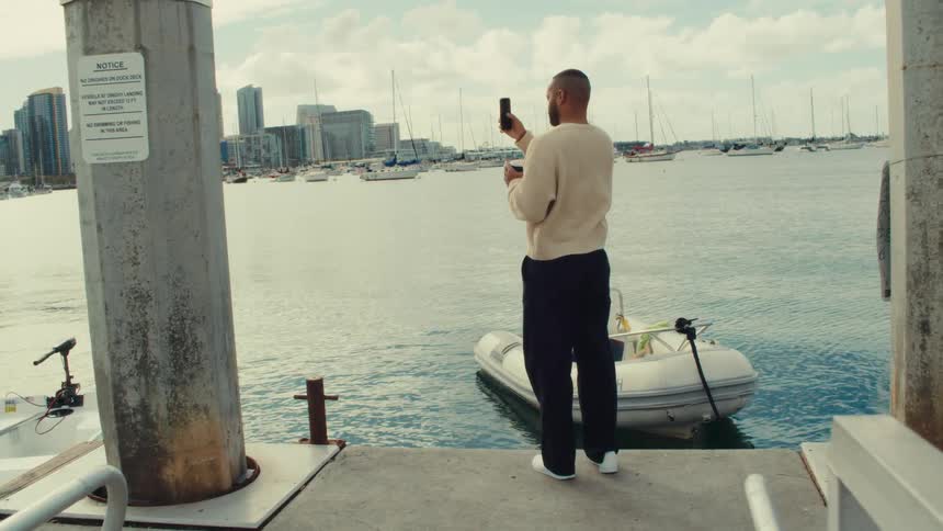 Man stands along the coast with boats on the water as he takes pictures with his phone