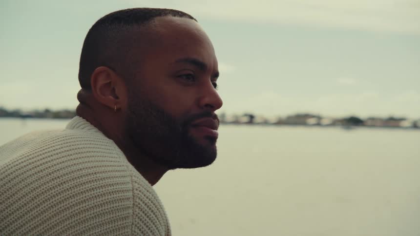 A man with a beard and gold earring wears a cream knit sweater while looking out over a calm body of water toward a distant shoreline under a bright sky