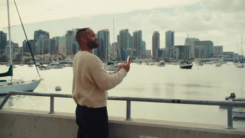 Man stands along the coast with boats in the harbor as he takes pictures with his phone