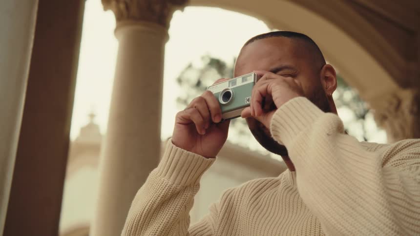 A man in a cream sweater walks into a courtyard and takes a picture with a small camera up to his eye while standing beneath an arched columned walkway