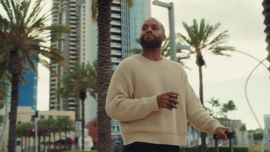 A medium shot shows a man wearing a cream sweater walking along a city sidewalk while holding a cup of coffee and pulling a suitcase past tall palm trees and modern buildings