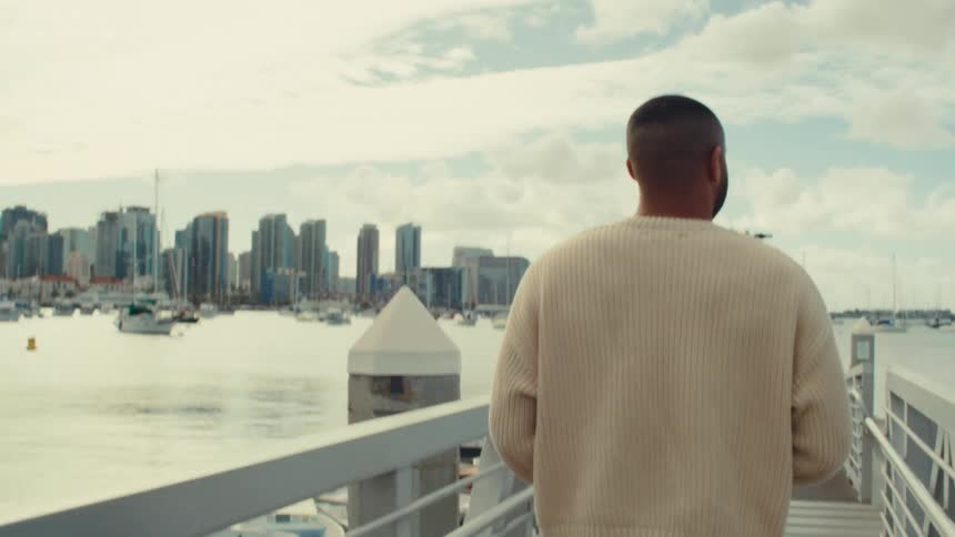 A black man walks on a waterfront pier looking out toward a city skyline across the harbor