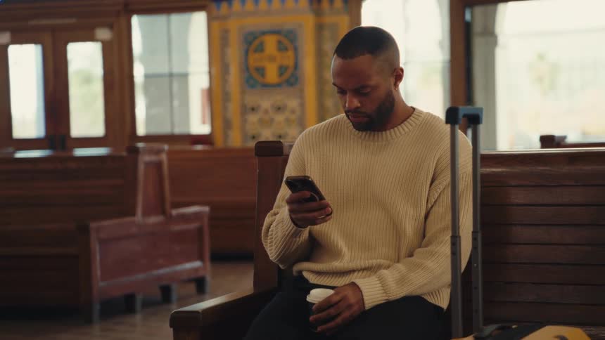A medium shot shows a man wearing a cream sweater as he sits on a wooden bench in a train station while looking at his phone and holding a cup of coffee next to a suitcase