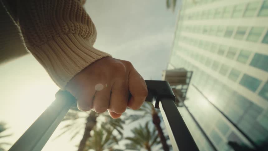 A low angle shot shows a hand in a cream sweater gripping a suitcase handle while walking past palm trees and a tall glass building under a bright sun