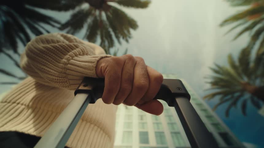 A low angle shot looking up shows a hand in a cream sweater grabbing a suitcase handle while walking outside under palm trees and a tall building against a bright sky
