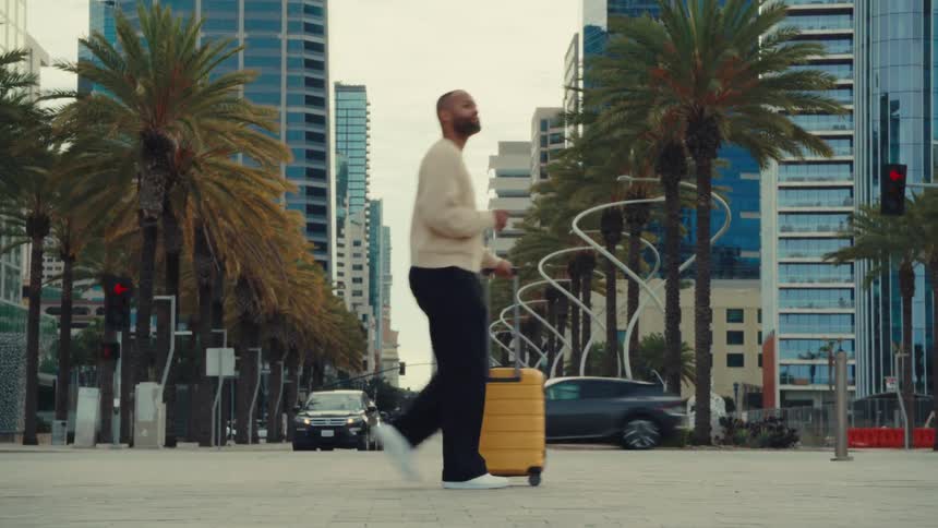 Man pulls a yellow suitcase while crossing an avenue with palm trees and tall buildings