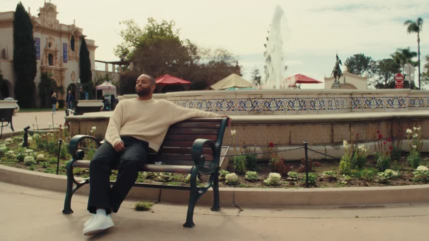 A man in a cream sweater relaxes on a park bench beside a decorative fountain in a lively plaza
