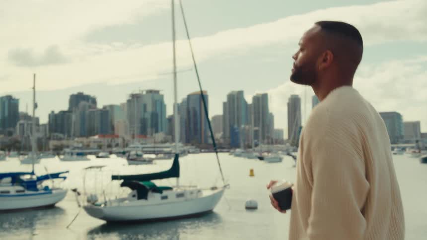 Man holds a coffee cup while looking at a marina with boats and buildings in the background