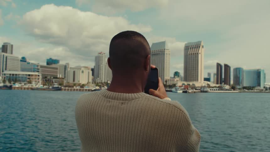 A man in a cream sweater takes video with a smart phone of a city skyline across the water from a waterfront viewpoint