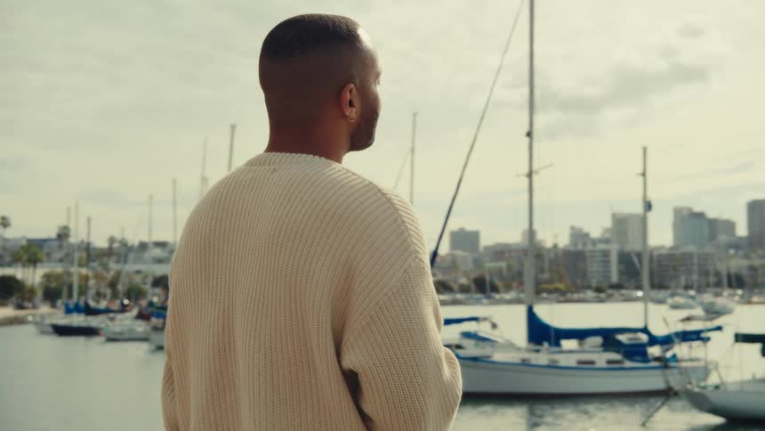 Person in beige sweater walks in marina with sailboats in background