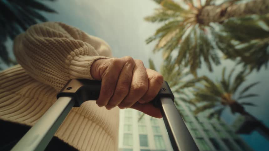 A closeup from a low angle shows a persons hand gripping a suitcase handle with palm trees and tall buildings in the background