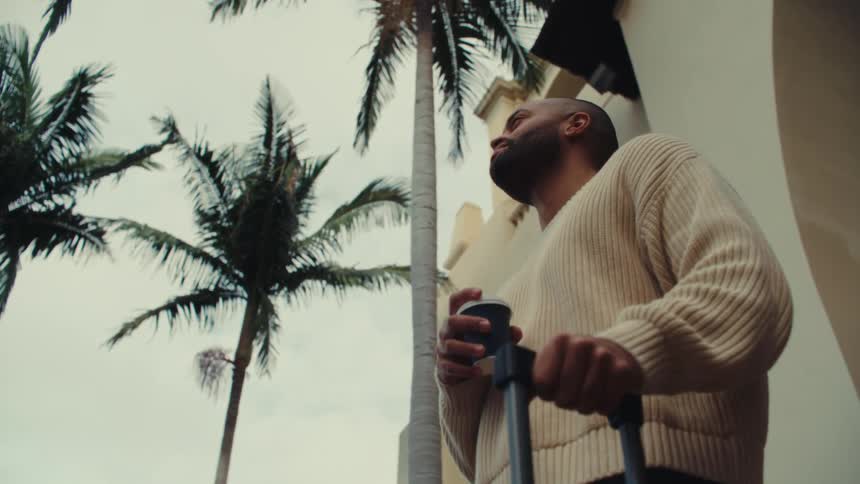 A low angle shot shows a man in a cream sweater holding a cup of coffee and the handle of a suitcase while looking up at tall palm trees and a light colored building under an overcast sky