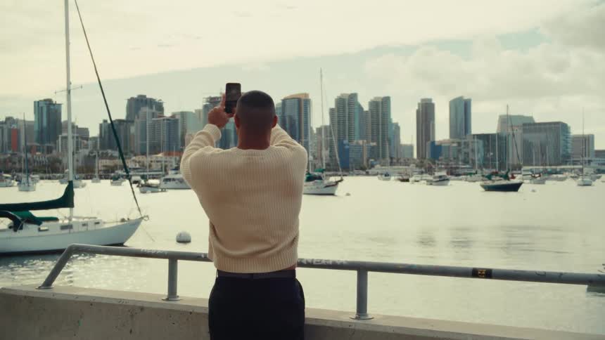 A shot from behind shows a man in a cream sweater as he stands at a railing and uses his phone to take a photo of a harbor filled with boats against a city skyline