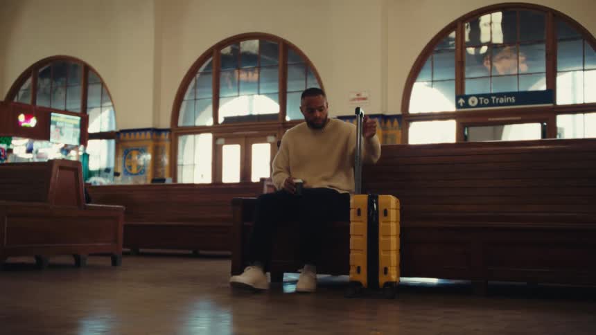 A medium shot shows a man in a cream sweater sitting on a wooden bench in a train station next to a yellow suitcase before he stands up and walks away while pulling the luggage