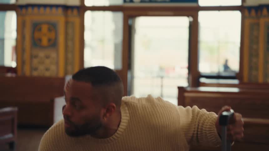 A medium shot shows a man in a cream sweater sitting on a wooden bench in a train station before he stands up and begins walking away while pulling a suitcase