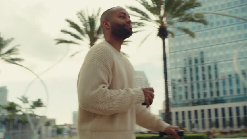 Person in beige sweater pulls suitcase and holds drink in urban area with palm trees