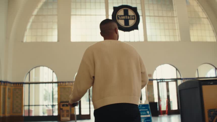 A shot from behind shows a man wearing a cream sweater as he walks through a spacious train station lobby toward arched glass doors while pulling a suitcase