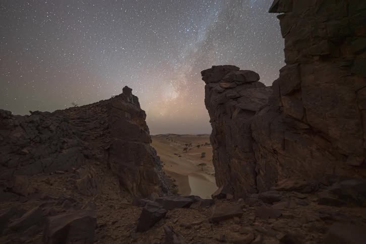 A time lapse view shows the milky way and countless stars moving across the night sky above dark jagged rock formations and sandy desert dunes as the sun sets and darkness falls over the remote landscape