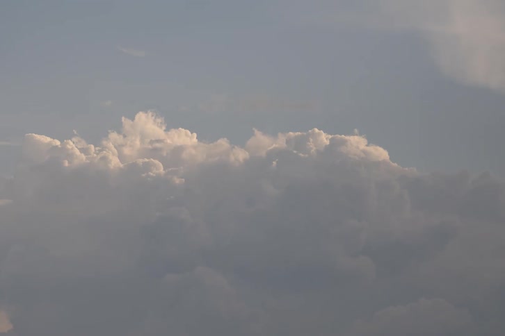 These are towering cumulus clouds glowing with soft golden sunlight against a muted sky the dense billowy formations shift slowly as they are partially eclipsed by a darker layer of foreground clouds