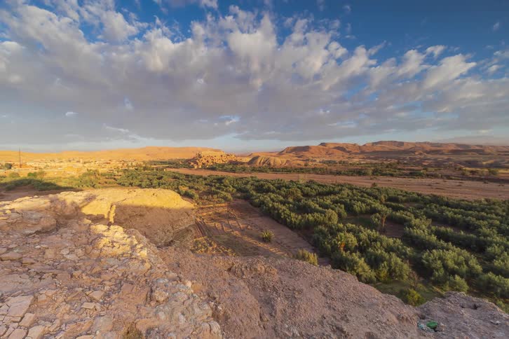 A time lapse view shows shadows stretching across a rocky desert overlook and a lush green valley filled with palm trees as clouds drift through a blue sky during sunset