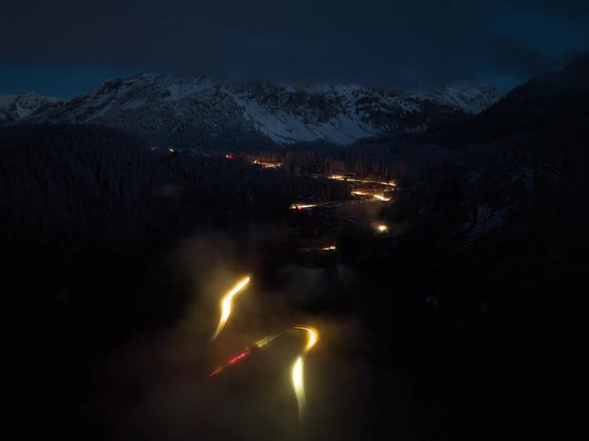 Headlights of automobiles glow as traffic moves on a winding road through forested mountains as darkness falls