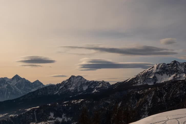 A time lapse view shows long flat clouds stretching across a pale sky above a range of snow covered mountains and dark pine forests during a cold winter evening