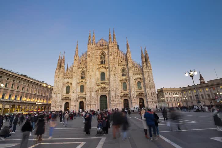 A large crowd of tourists gathers in the plaza in front of the historic milan cathedral at dusk