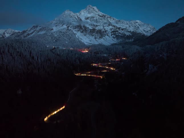 Headlights of automobiles glow as traffic moves on a winding road through forested mountains as darkness falls