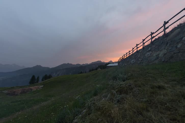 A time lapse view shows a golden sunset fading behind distant mountains while shadows grow over a grassy hillside and a stone wall with a wooden fence during a quiet evening