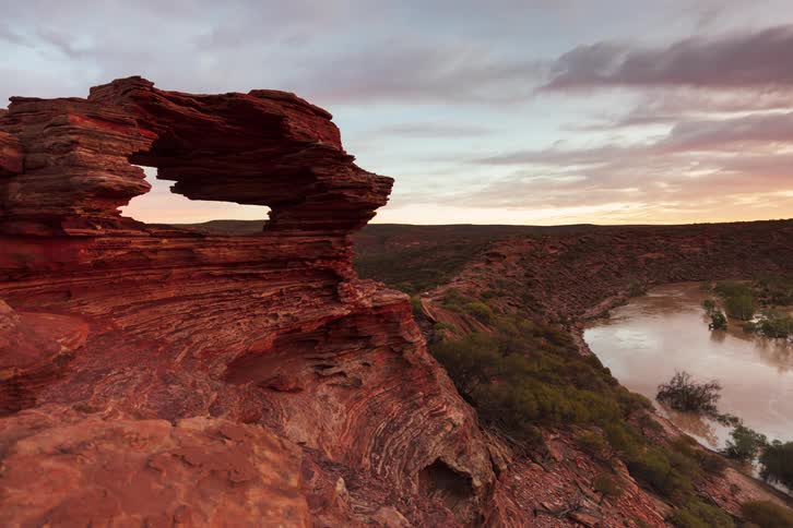 A natural rock arch overlooks a winding river mountains and a rugged canyon landscape under a cloudy sky