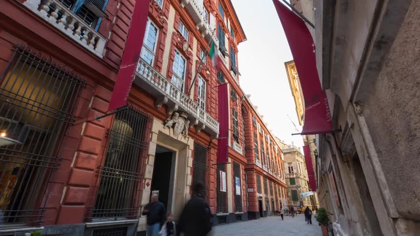 A time lapse view shows people walking along a narrow paved street past a grand orange building with red banners and ornate stone carvings on a bright day