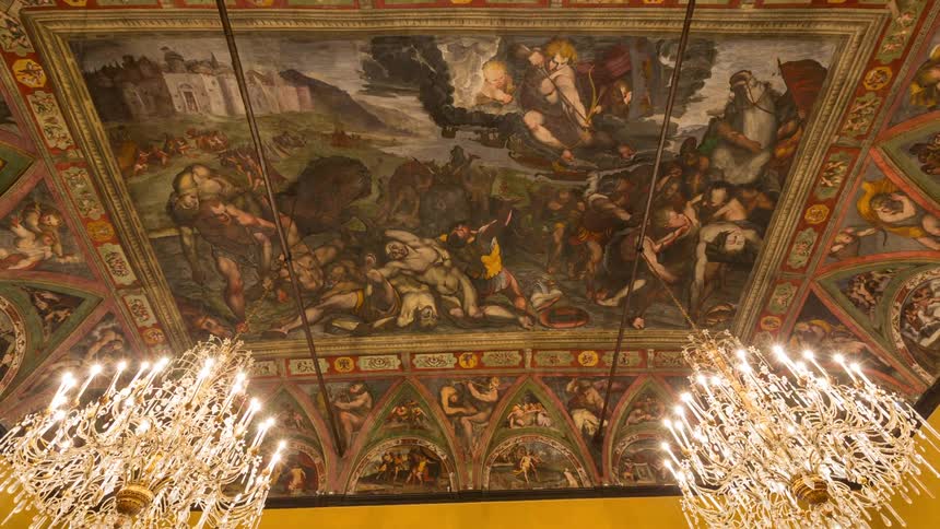 A vast renaissance fresco depicts a dramatic mythological battle across a grand ceiling framed by two glowing crystal chandeliers and intricate decorative borders in a historic palatial hall