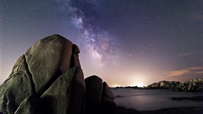 Shooting stars pass through the dark sky at sunrise over a boulder strewn coast