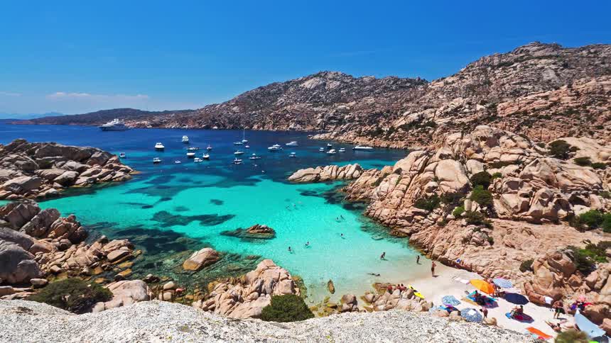 A beach surrounded by rocks as people walk in and out of the water on a sunny day