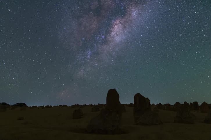 The milky way spans across a starfilled night sky above rock formations in a desert landscape