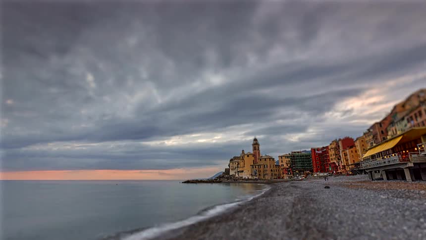 A coastal town with colorful buildings and a church tower sits along a calm shoreline with a few beachgoers under a dramatic cloudy sky