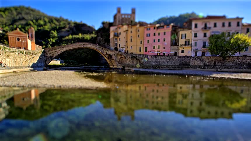 The video showcases a picturesque stone arch bridge that spans a calm river connecting two sides of a historic villagealong the riverbank a row of colorful multistory buildings is topped by an ancient stone castle or fortress perched o