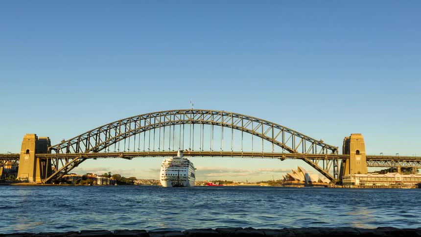 A large white cruise ship glides through a harbor passing directly beneath the massive steel arch of the sydney harbour bridgethe scene captures the iconic bridge and a glimpse of the sydney opera house under a clear bright sky