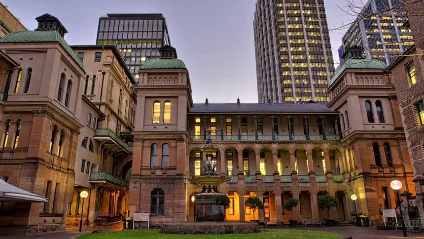 A historic building with arched balconies and green domes stands in a courtyard withpeople walking and a statue fountain surrounded by modern skyscrapers