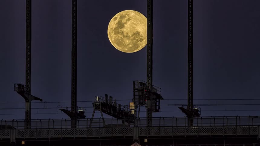 Full moon above industrial structures and power lines against a dark night sky
