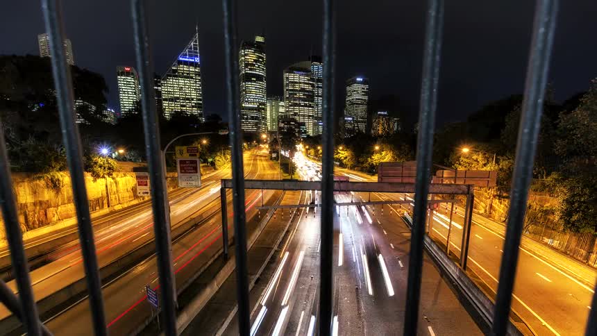 Metal bars in the foreground facing a highway with vehicle light trails and illuminated buildings at night