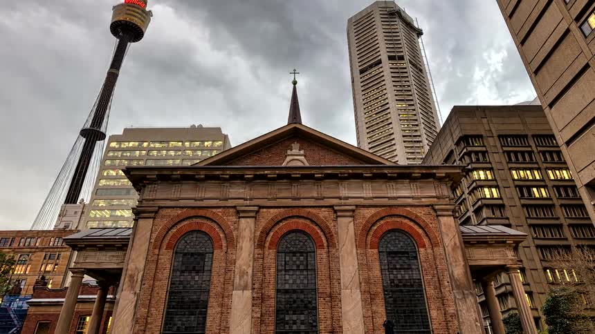 A time lapse view shows a historic stone church surrounded by modern skyscrapers and an observation tower as the grey cloudy sky darkens and warm lights illuminate the building facade