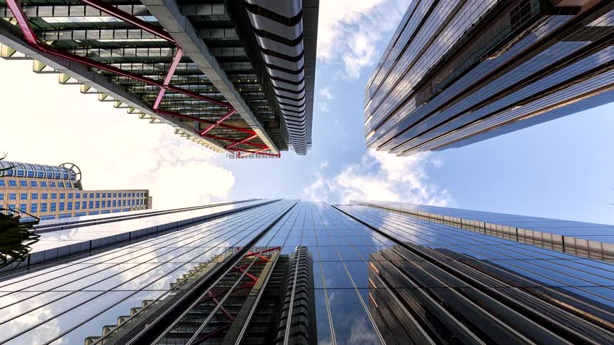 A low view looking up at modern glass and steel skyscrapers reaching toward a blue sky with soft white clouds reflecting in the windows of the tall buildings