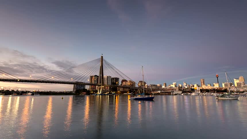 A time lapse view shows a suspension bridge and a city skyline reflecting in the water as pink sunset clouds fade into a dark night sky and city lights begin to glow across the harbor