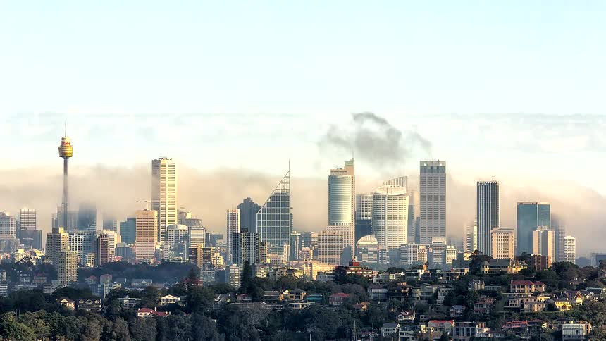 The video shows a wide shot of the sydney skyline as thick fog rolls through the buildings under a bright morning skylowlying clouds partially obscure the skyscrapers with the sydney tower eye visible on the left and residential areas in the foregrou